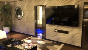 Uma sala de estar moderna com uma grande televisão de tela plana na parede, um vaso de planta, uma mesa de centro de vidro com revistas e um tapete estampado.