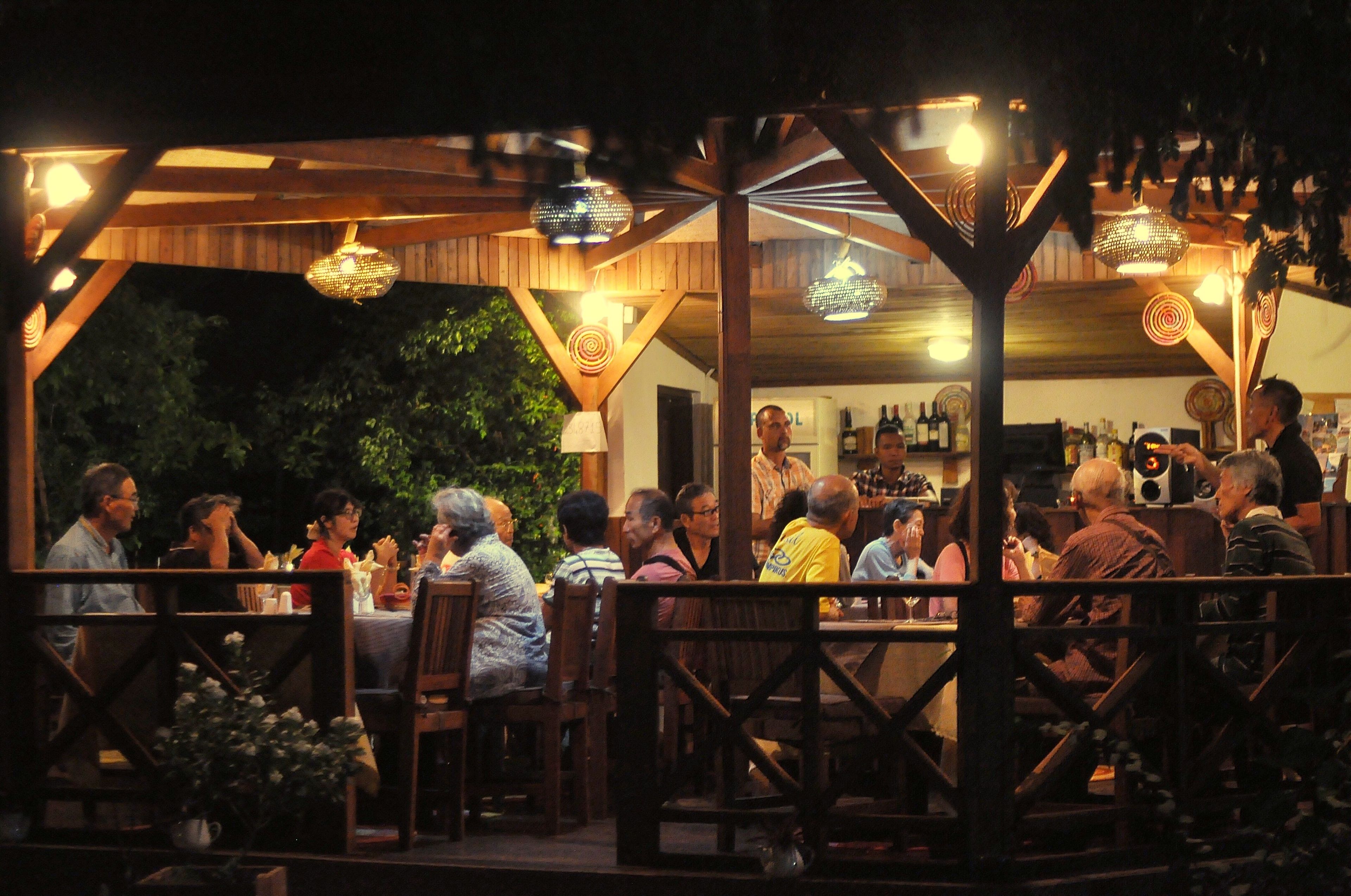An outdoor dining area with wooden tables and chairs, under a covered structure with hanging lights.