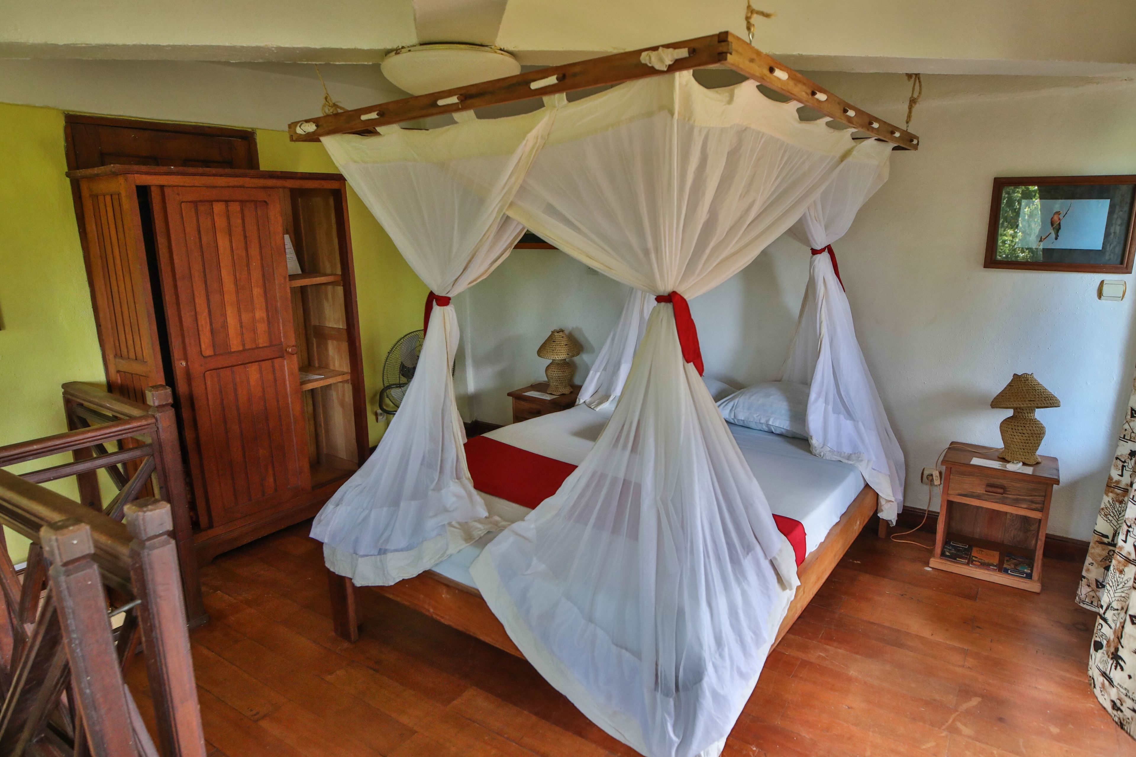 A room with a bed under a canopy, a wooden wardrobe, a bedside table with a lamp, and a small shelf with books.