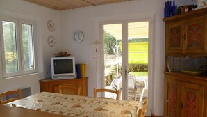 Un coin repas avec une table et des chaises, un téléviseur posé sur un meuble en bois, et une vue sur l’extérieur grâce à une porte vitrée.