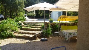 Un espace patio avec une table et des chaises sous une pergola, entouré d’un jardin et d’un mur de pierre.