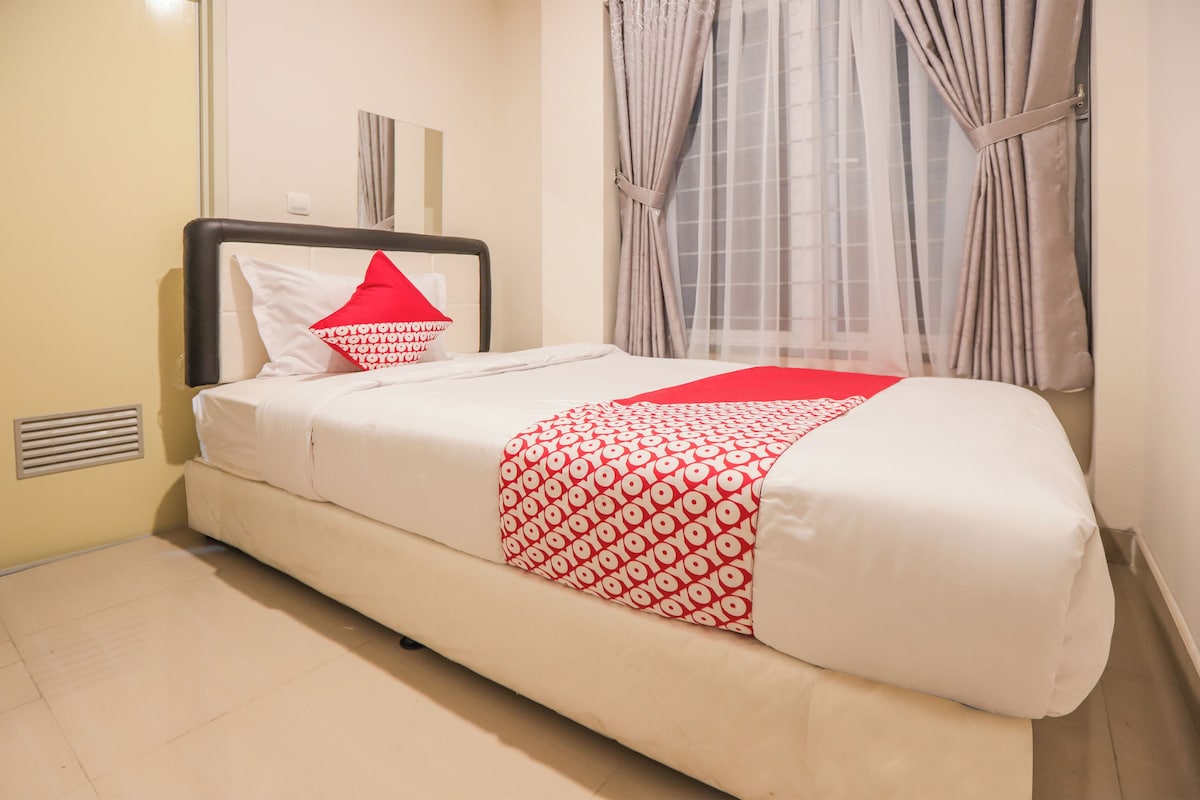 Un lit bien fait, recouvert d’une couvre-lit à motifs blancs et rouges, avec une tête de lit noire et un seul oreiller rouge.