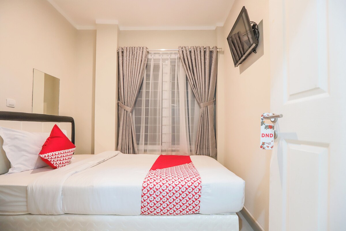 Una cama bien hecha con ropa blanca y un corredor rojo y blanco estampado. Hay un cojín rojo sobre la cama, y un televisor montado en la pared encima.