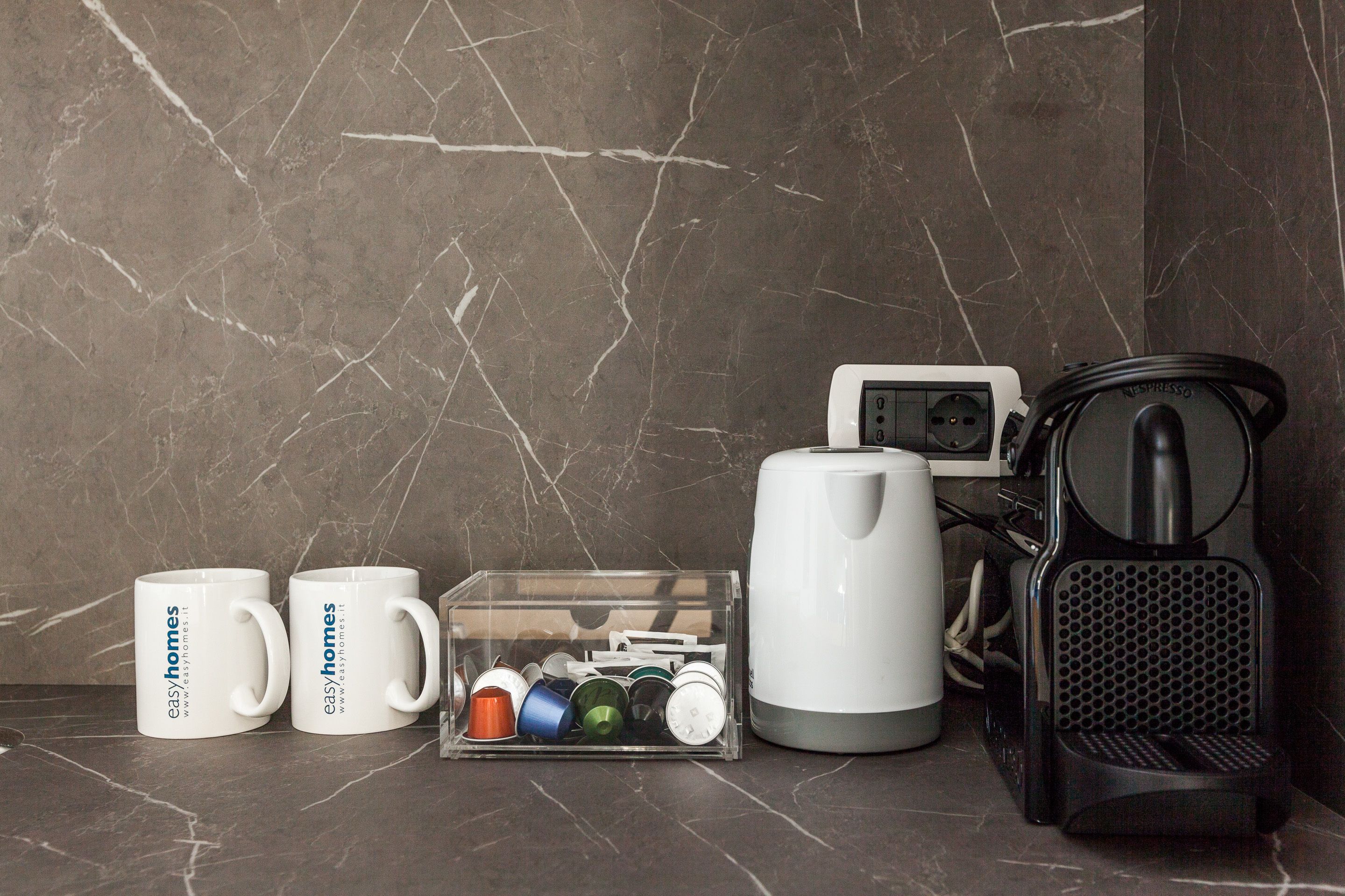 A kitchen counter with a coffee maker, two mugs, a clear storage box with coffee pods, and a kettle.