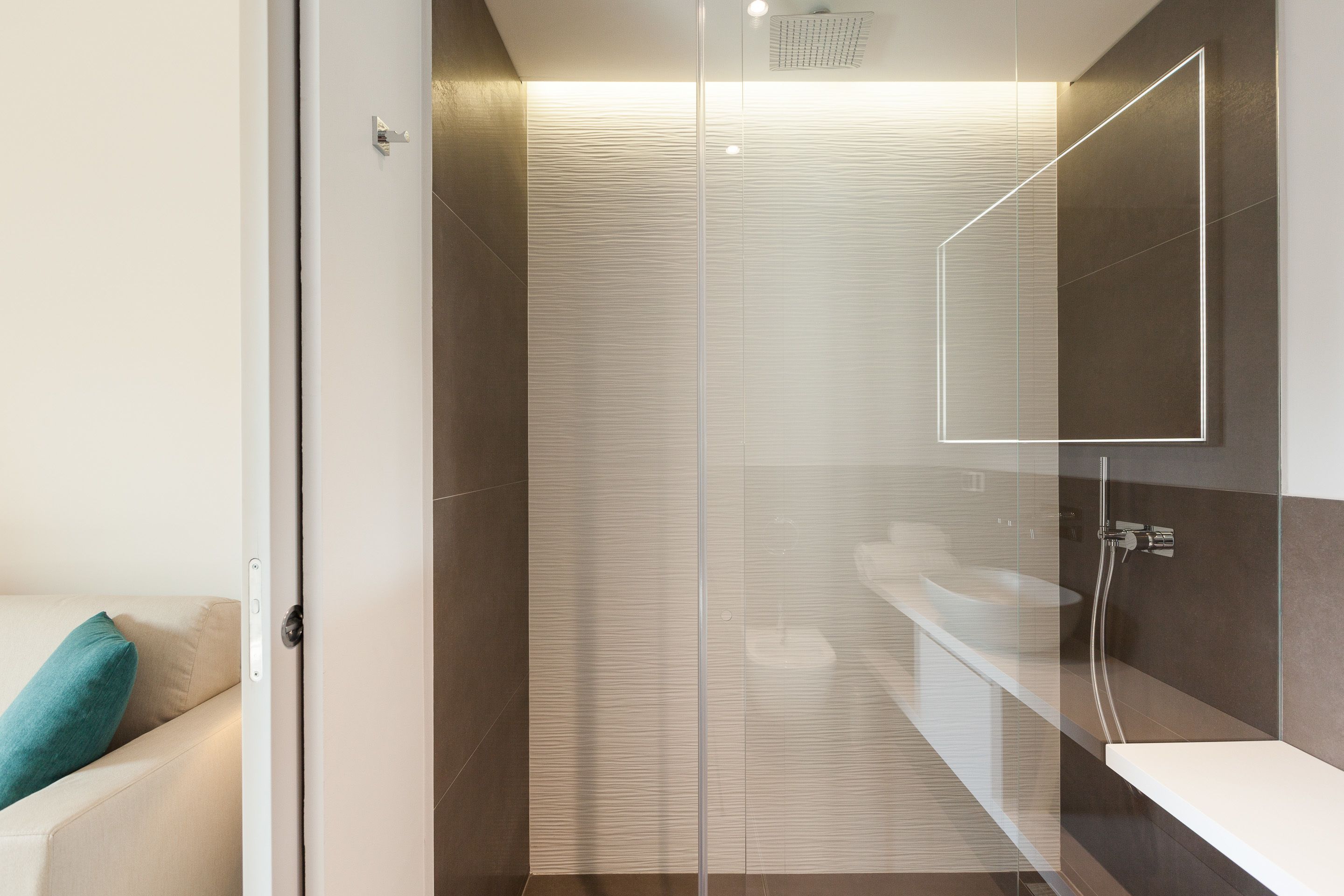 A modern bathroom with a glass shower enclosure, a white sink, and a beige sofa with a teal pillow.