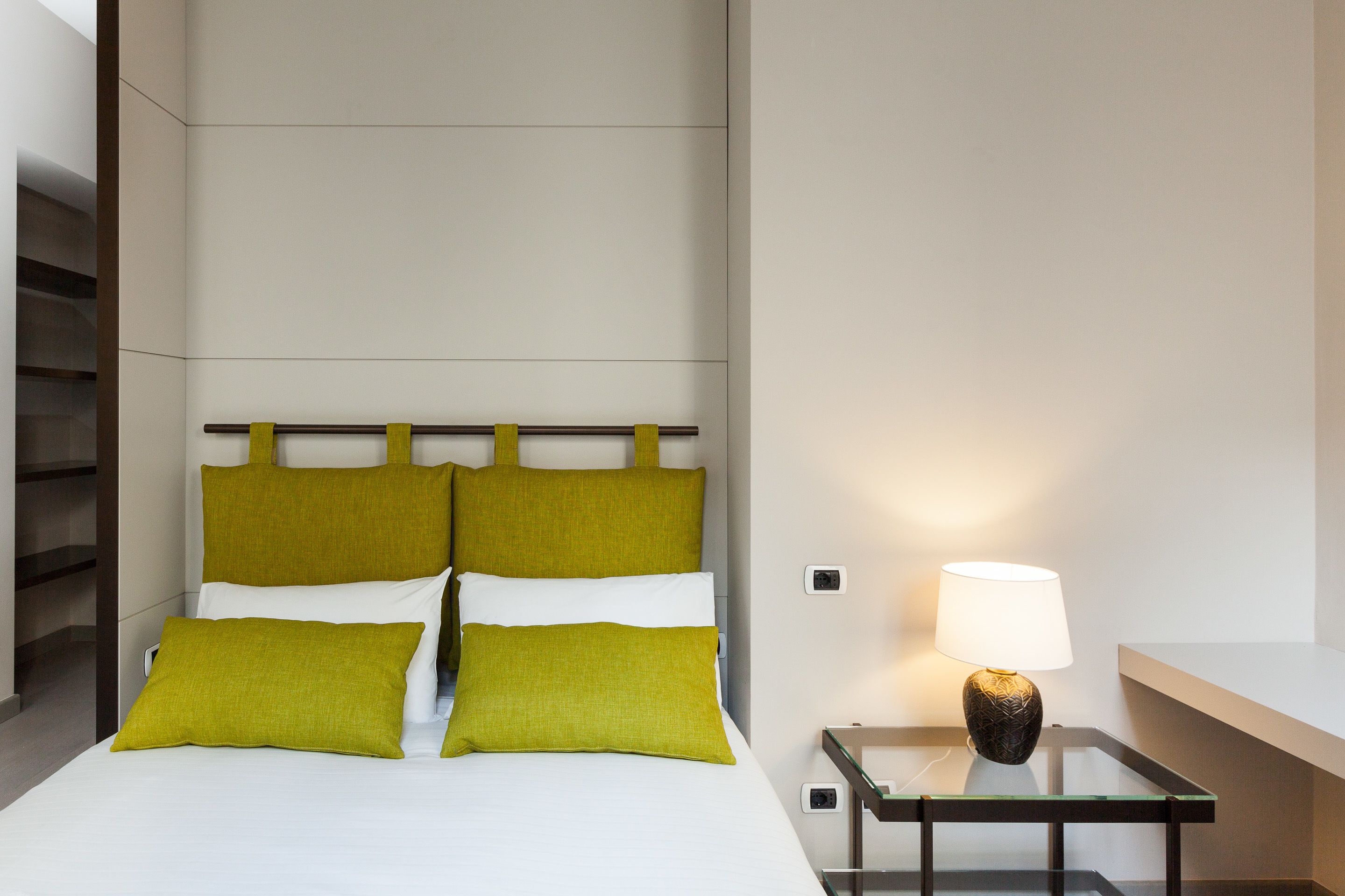 A hotel room with a bed, green pillows, a bedside table with a lamp, and a glass partition.