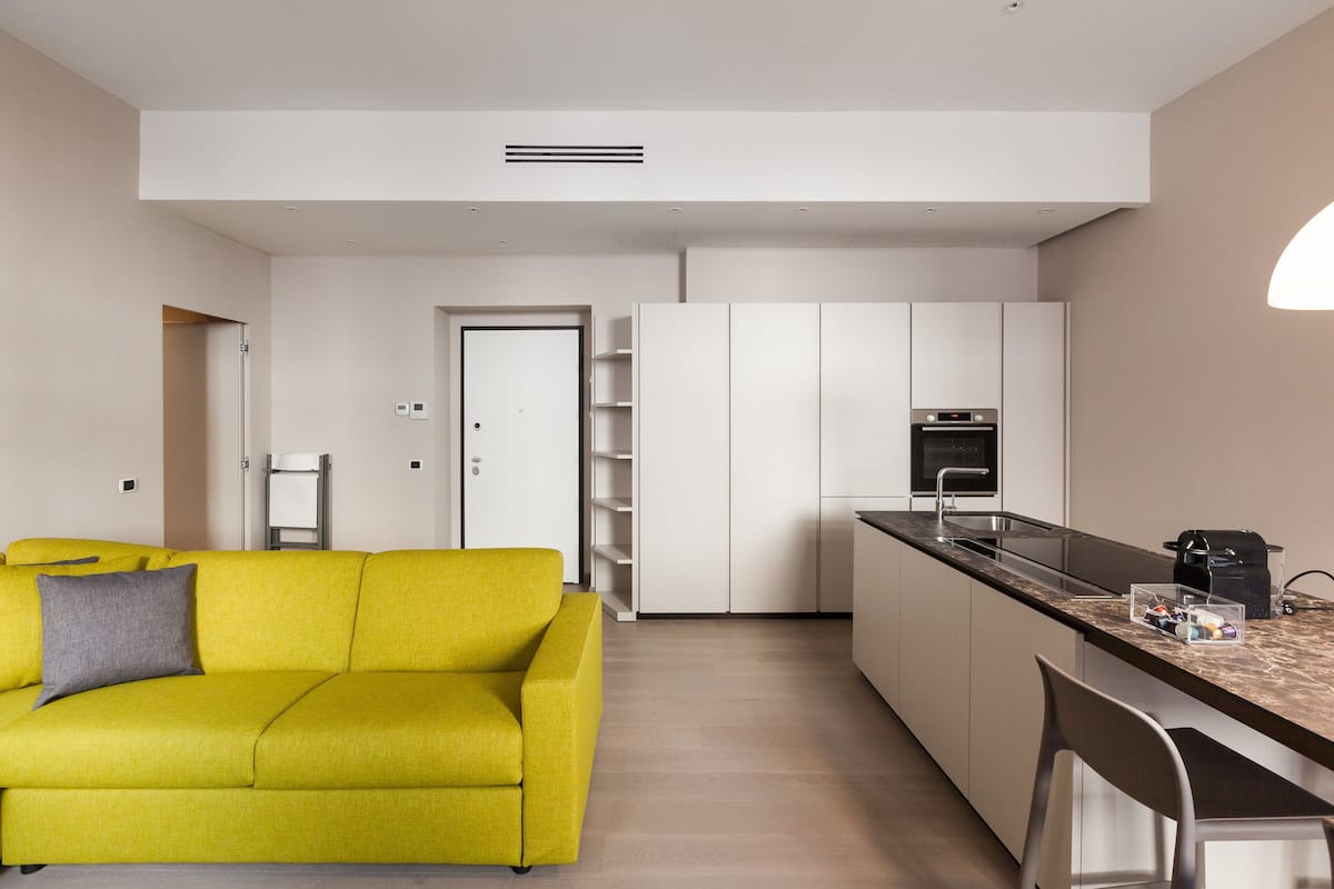 A modern apartment with a bright yellow sofa, a kitchen with white cabinets, and a marble countertop.