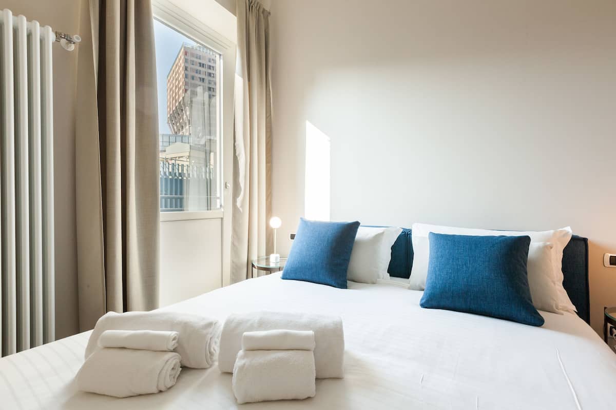A hotel room with a large bed, white linens, and blue pillows. There are towels on the bed and a window with curtains.
