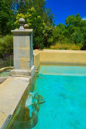 Une piscine à l’eau d’un bleu cristallin, entourée de verdure et bordée d’une structure en pierre surmontée d’une sphère.