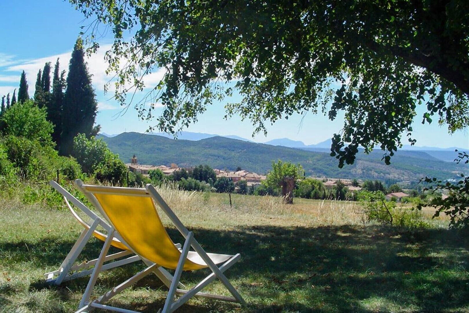 Une chaise jaune placée sous un arbre, d’où l’on peut admirer un paysage rural avec des montagnes à l’horizon.