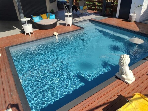 Una piscina con cubierta de madera, una estatua blanca y un área de descanso con cojines y una mesita.