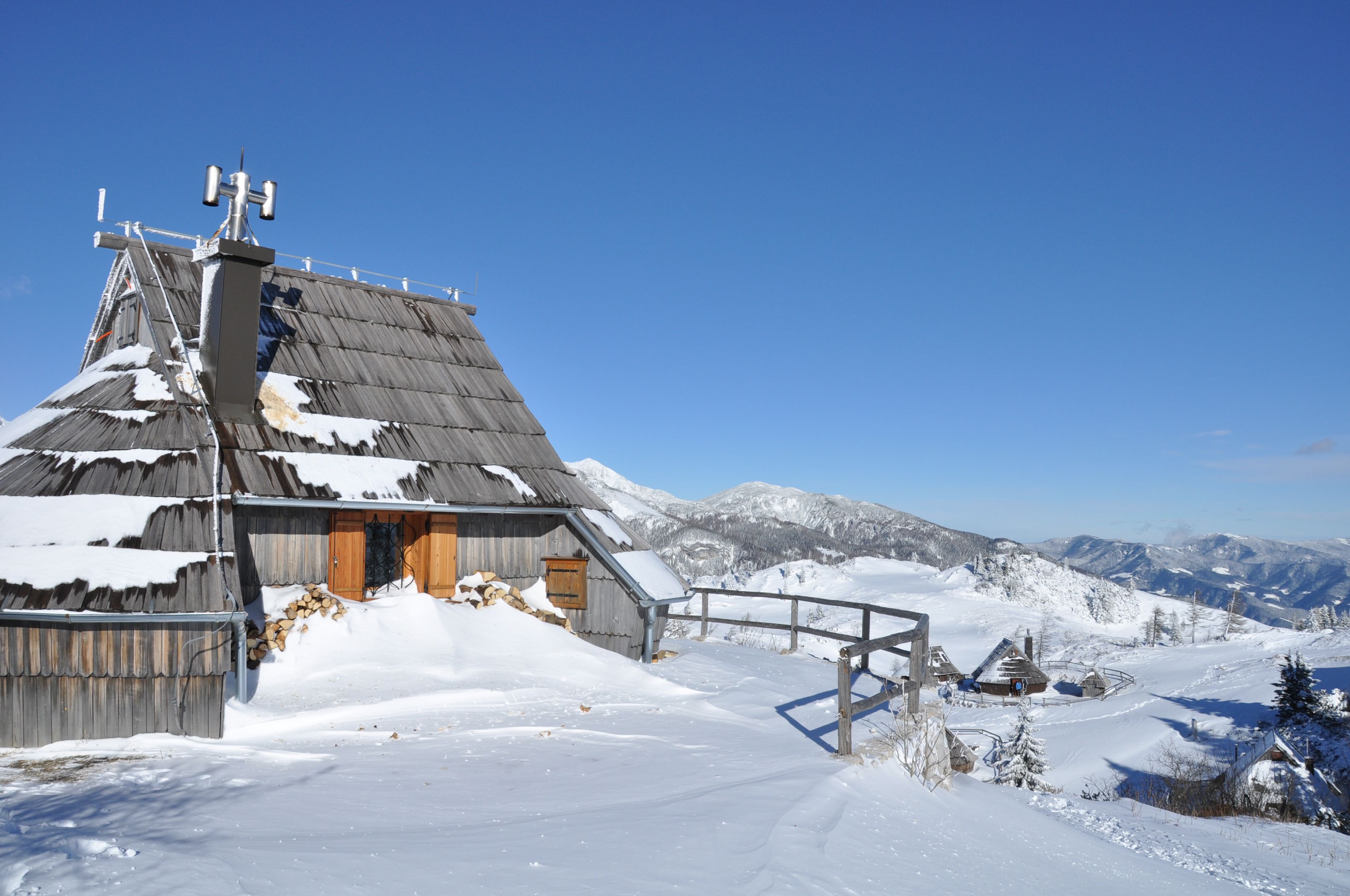 A wooden cabin with a sloped roof, surrounded by snow-covered mountains and a clear blue sky.