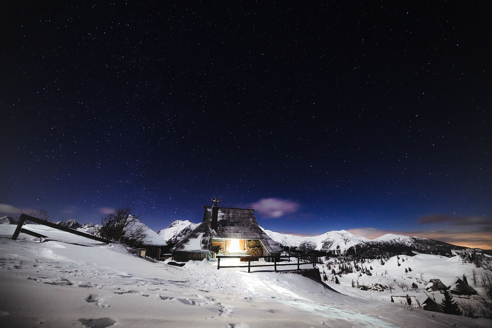 A snow-covered mountain landscape at night with a lit building in the center, surrounded by snow and a clear sky.