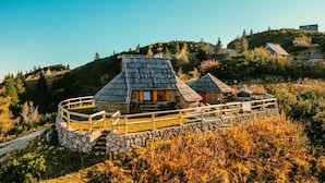 A traditional wooden house with a stone foundation and a wooden fence, surrounded by a natural landscape with trees and hills. - Koca Ojstrica - Velika planina (Kamnik)