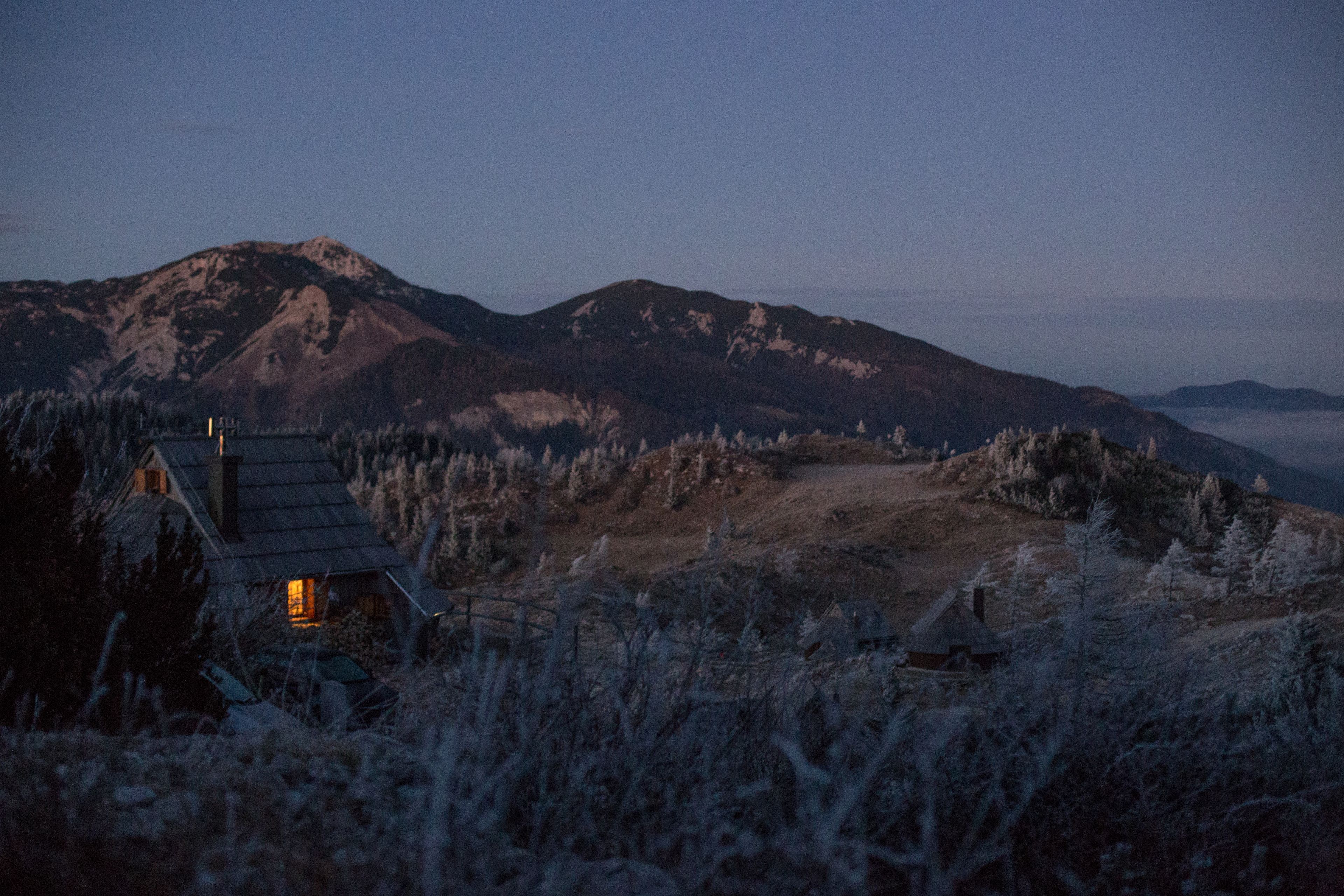 A rustic cabin with a sloped roof, surrounded by a forested landscape and mountains in the background.