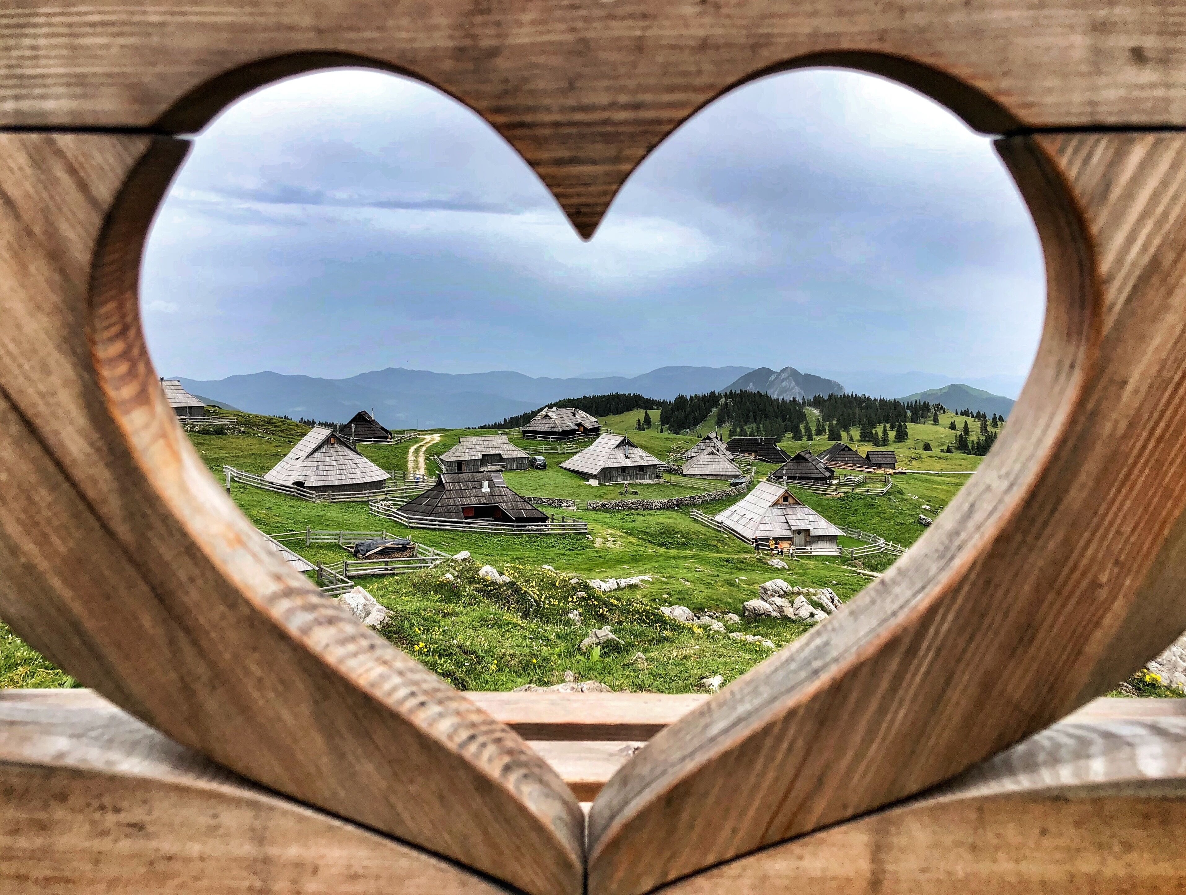 Un marco de madera con forma de corazón mostrando una vista de casas de madera tradicionales en un paisaje rural.