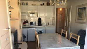 Een compacte keuken met witte kastjes, een magnetron, een koffiezetapparaat en een wasmachine. Er is een eettafel met stoelen en een kroonluchter.