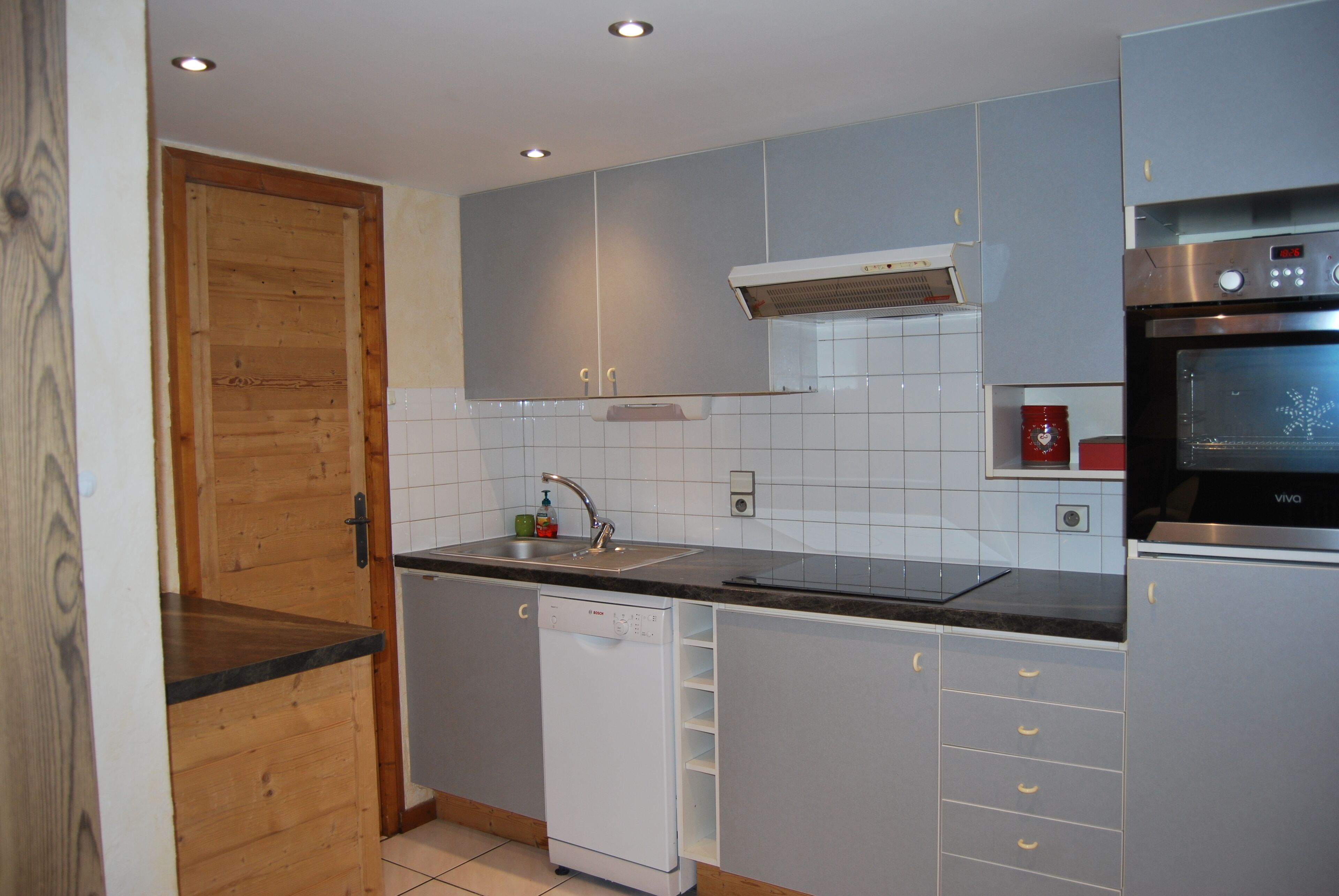 Una cocina moderna con gabinetes grises, lavavajillas, horno empotrado y puerta de madera.