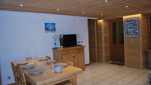 Un coin repas avec une table en bois dressée pour quatre personnes, un téléviseur posé sur un meuble en bois, et une porte donnant sur une autre pièce.