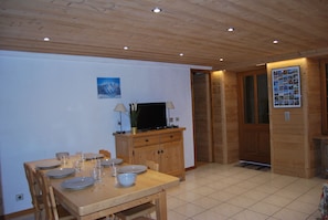 Een eetgedeelte met een houten tafel gedekt voor vier personen, een televisie op een houten kast, en een deur die naar een andere kamer leidt.