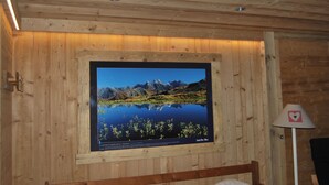 L’intérieur d’un chalet en bois, avec une photographie encadrée représentant un paysage montagneux accrochée au mur, un lit et une lampe.