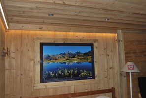 Een houten blokhut van binnen met een ingelijst foto van een berglandschap aan de muur, een bed en een lamp.