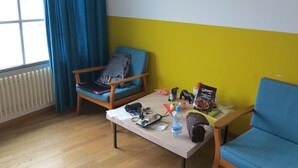 A room with a wooden floor, a blue chair, a table with various items, a radiator, and a window with blue curtains. - Basel Backpack - Hostel (Basel)