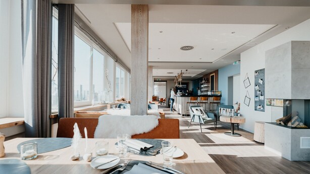 Un moderno comedor con techo alto, ventanas amplias y vistas al horizonte de la ciudad. Se observa una zona de bar al fondo con una barra y taburetes.