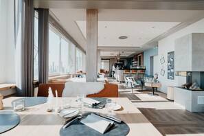 Uma zona de refeições moderna com teto alto, janelas amplas e vista para o skyline da cidade. Dá para ver uma área de bar ao fundo, com balcão e bancos.