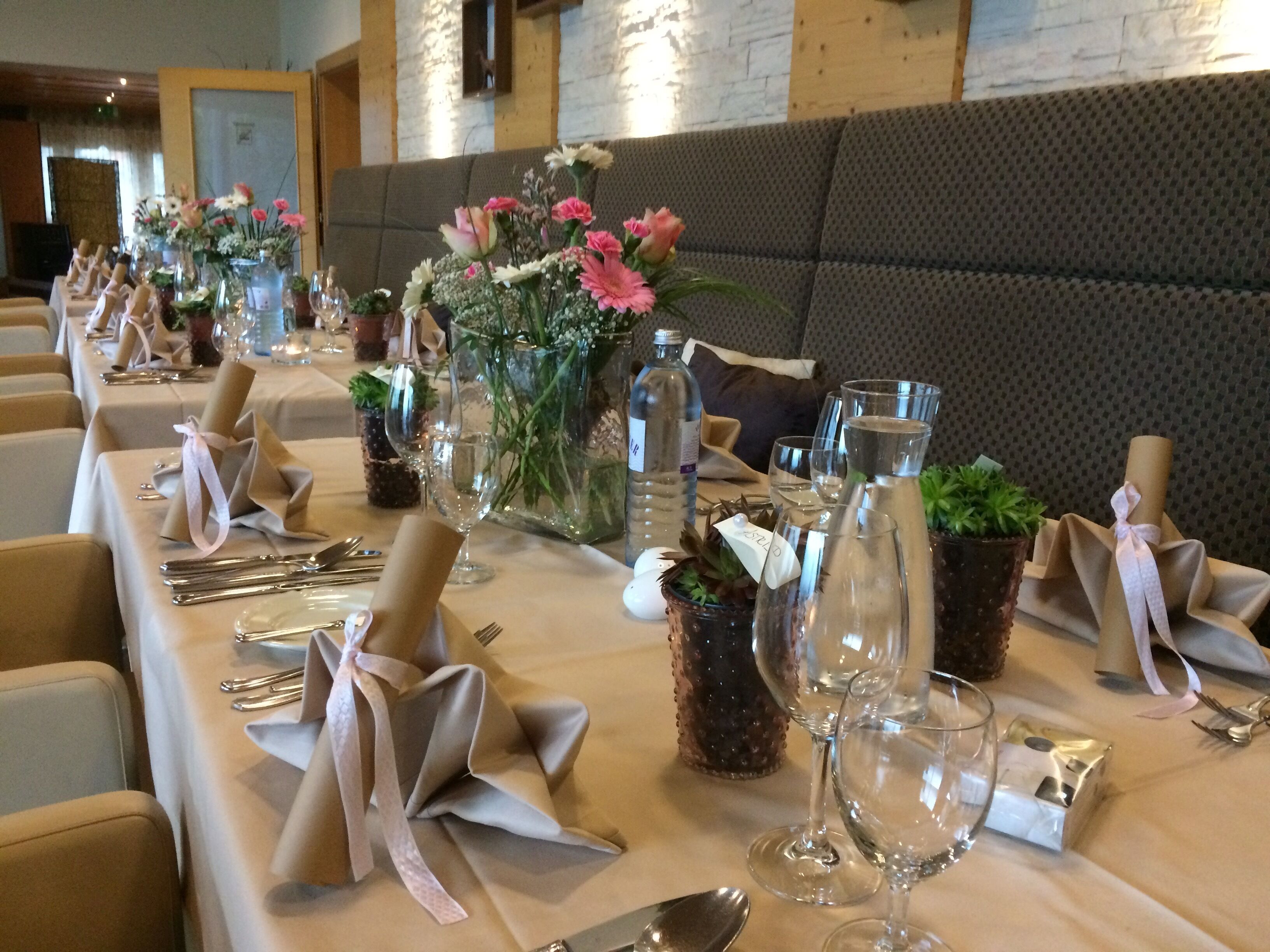 A banquet table set with glassware, plates, and napkins tied with ribbons, with floral centerpieces and a potted plant.