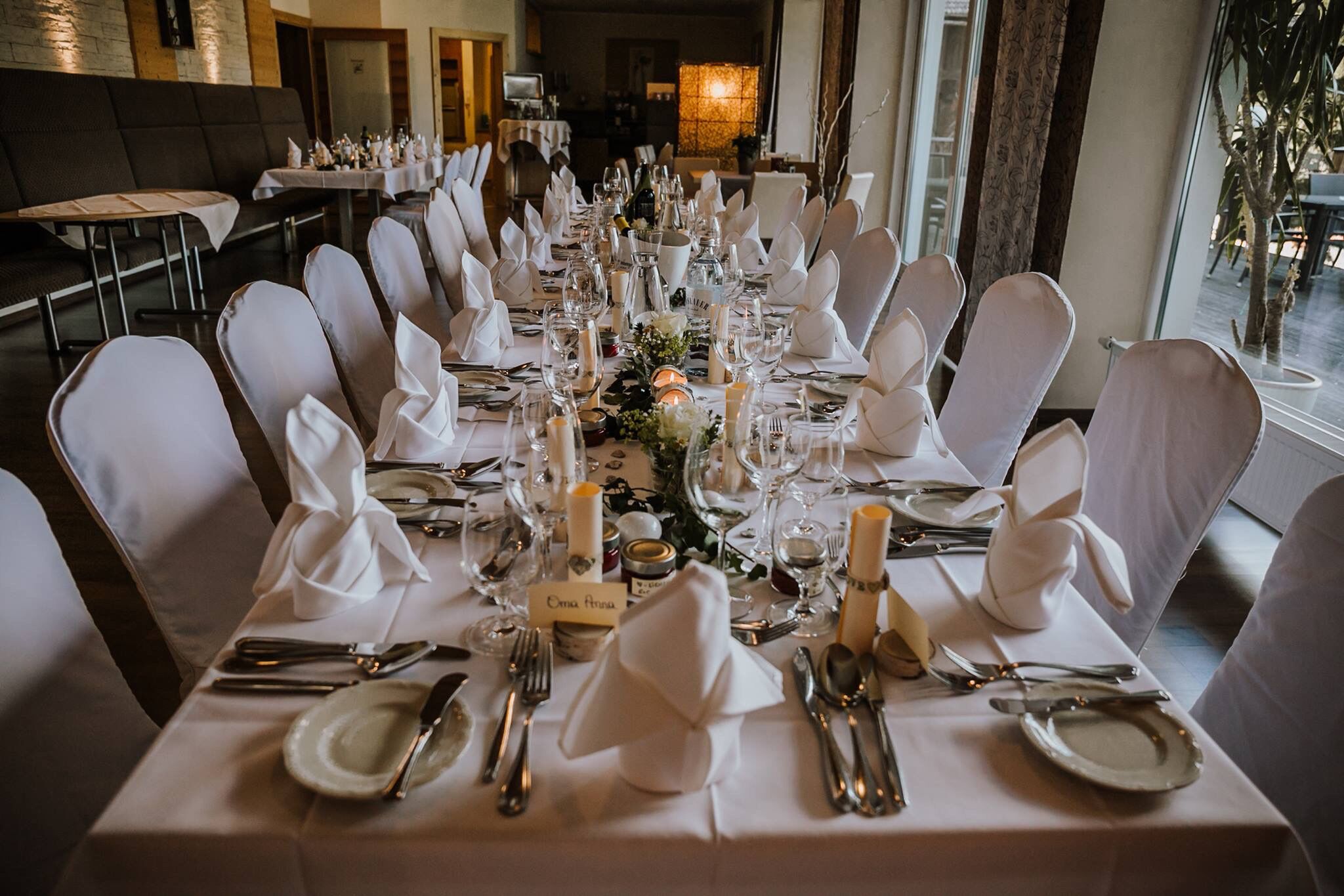 Una larga mesa de banquete dispuesta para un evento formal, con manteles blancos, velas y cristalería.