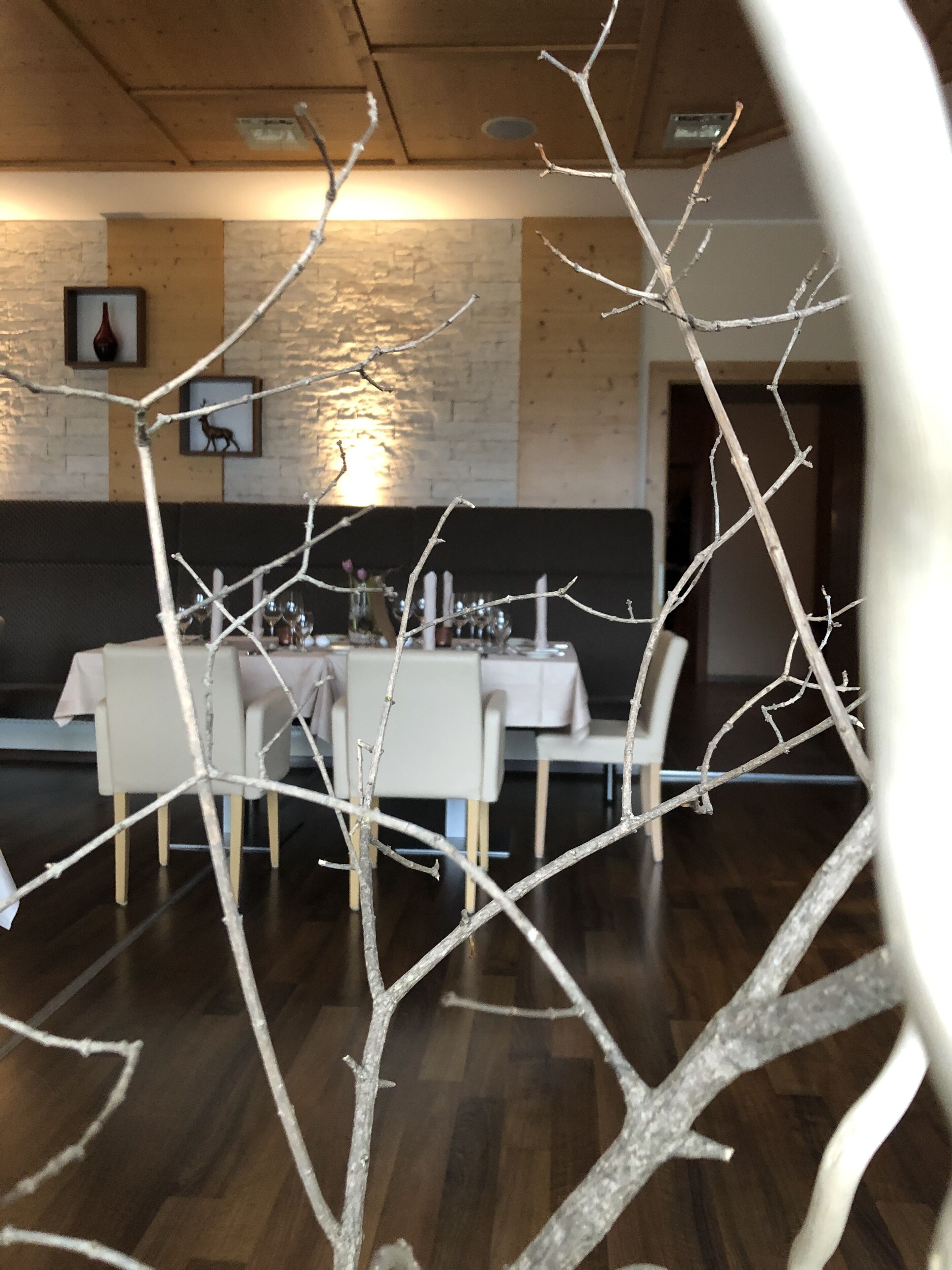 A dining area with a wooden table set for four, surrounded by chairs. A brick wall and a framed picture are visible in the background.