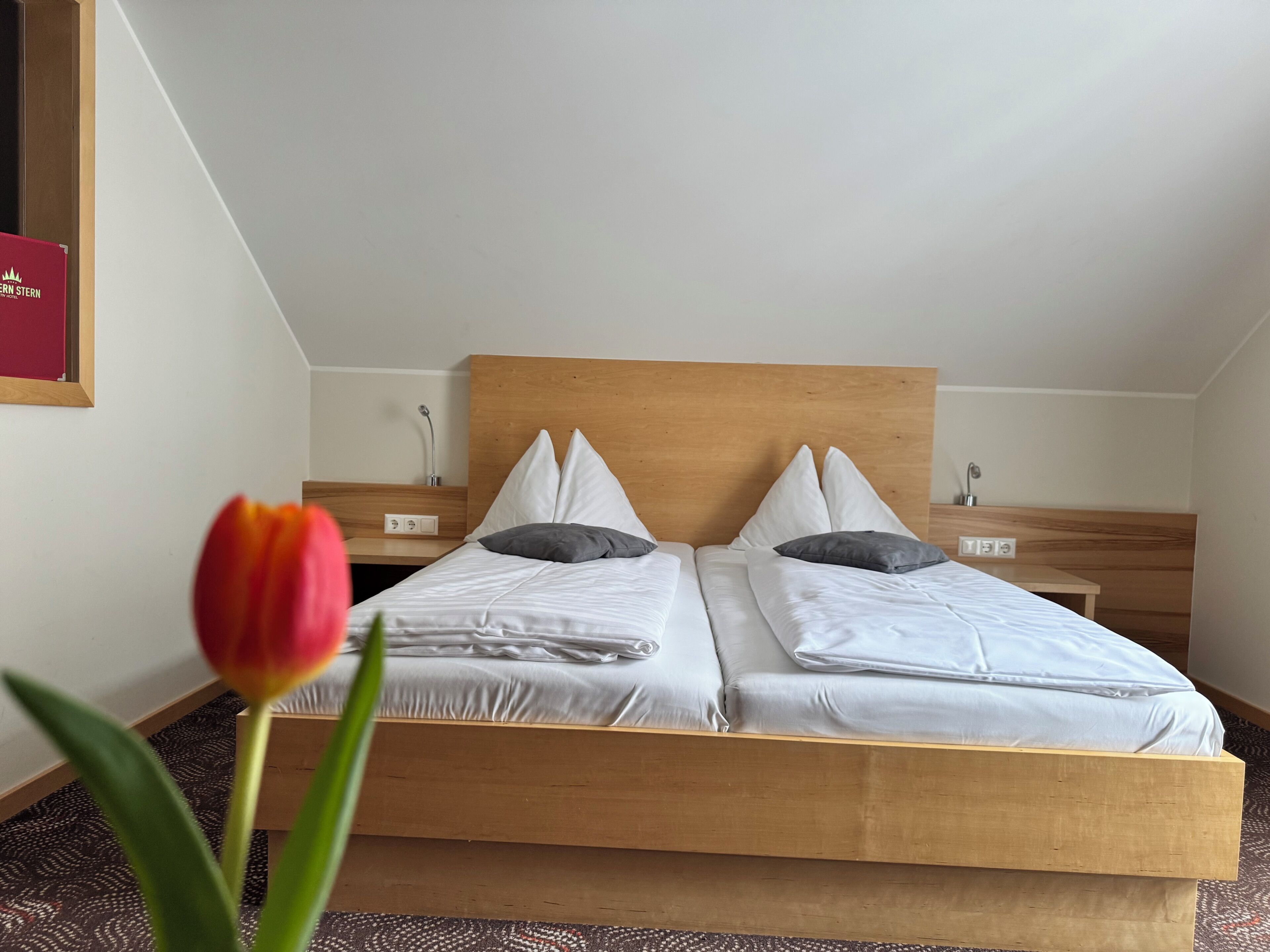 Una cama doble con cabecera de madera, dos mesitas de noche y una sola tulipán roja en primer plano.
