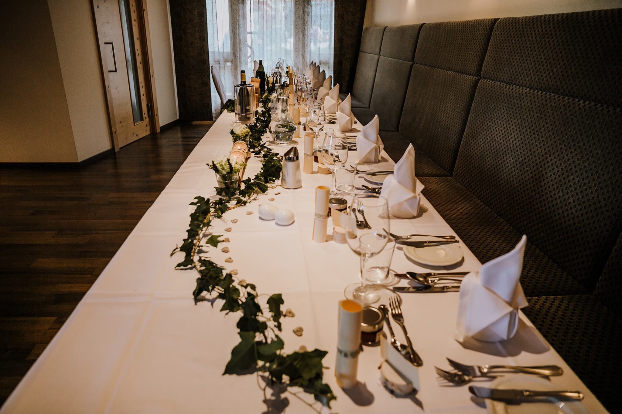 Una larga mesa de comedor vestida con mantel blanco, velas y follaje, en una habitación con sofá de cuero oscuro y piso de madera.