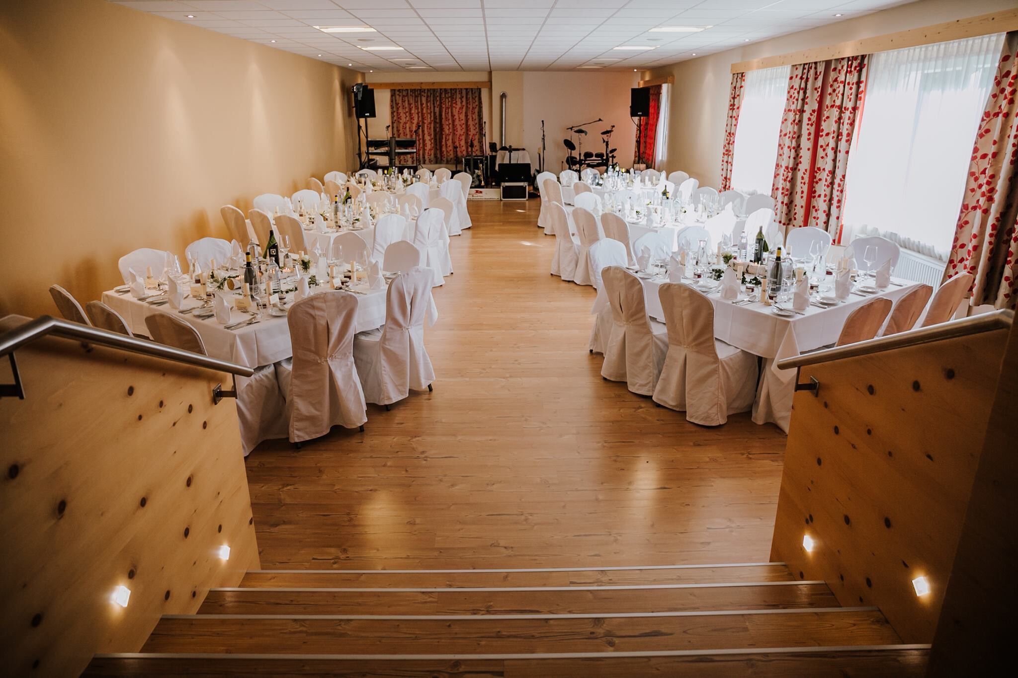 A formal dining hall with rows of tables set for a meal, a stage with musical instruments, and a staircase with wooden railings.