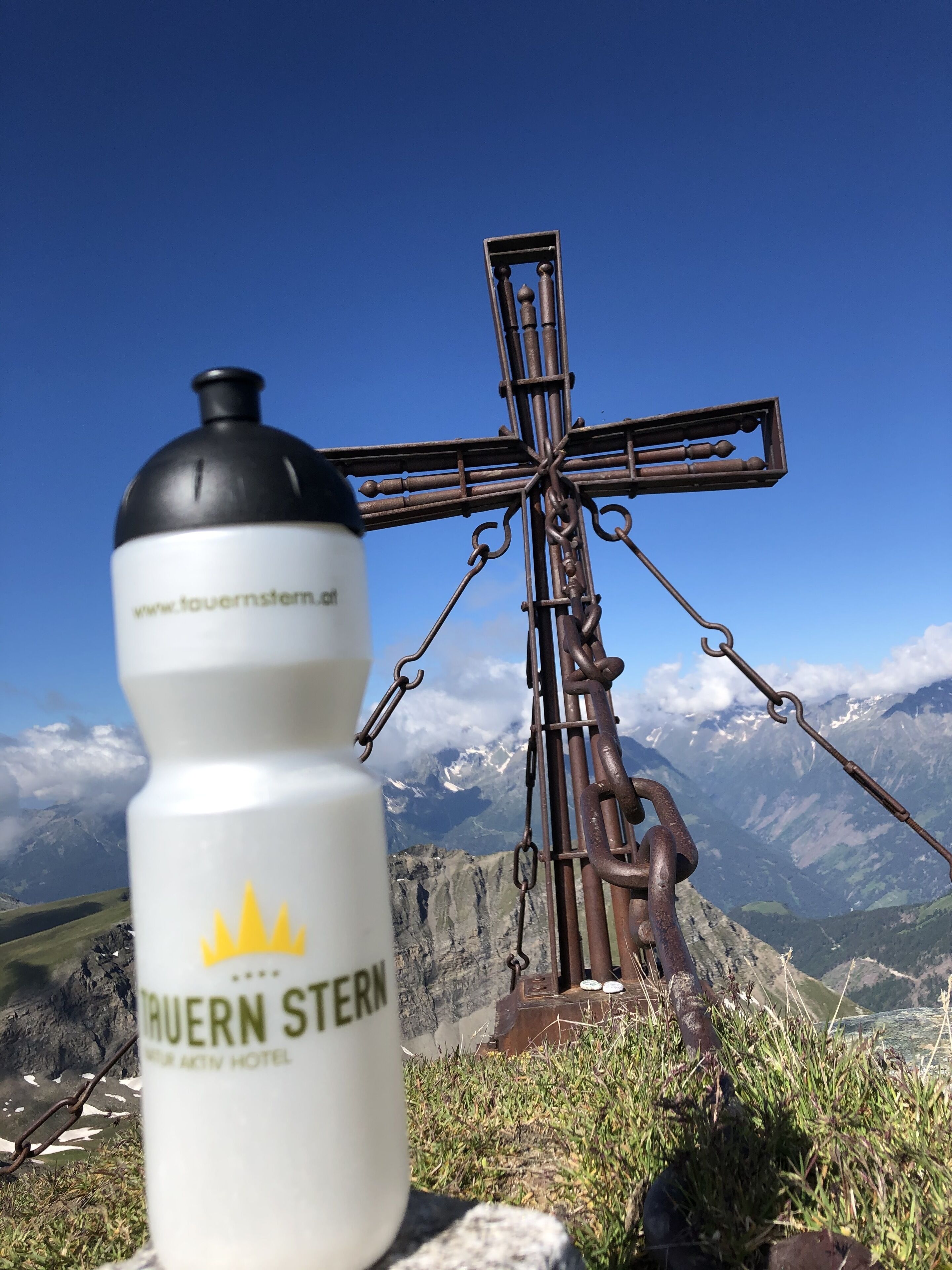 A bottle with "TRUERN STERN" and "www.touernstern.at" on it, in front of a metal cross structure with a mountainous backdrop.