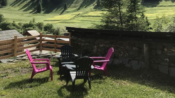 Une terrasse en bois avec des sièges extérieurs et un mur en pierre, offrant une vue sur un paysage montagneux avec de vastes forêts et un ciel dégagé.