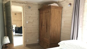 Une chambre avec une armoire en bois, un lit, une table de chevet avec une lampe, et une vue sur le salon par une porte ouverte.