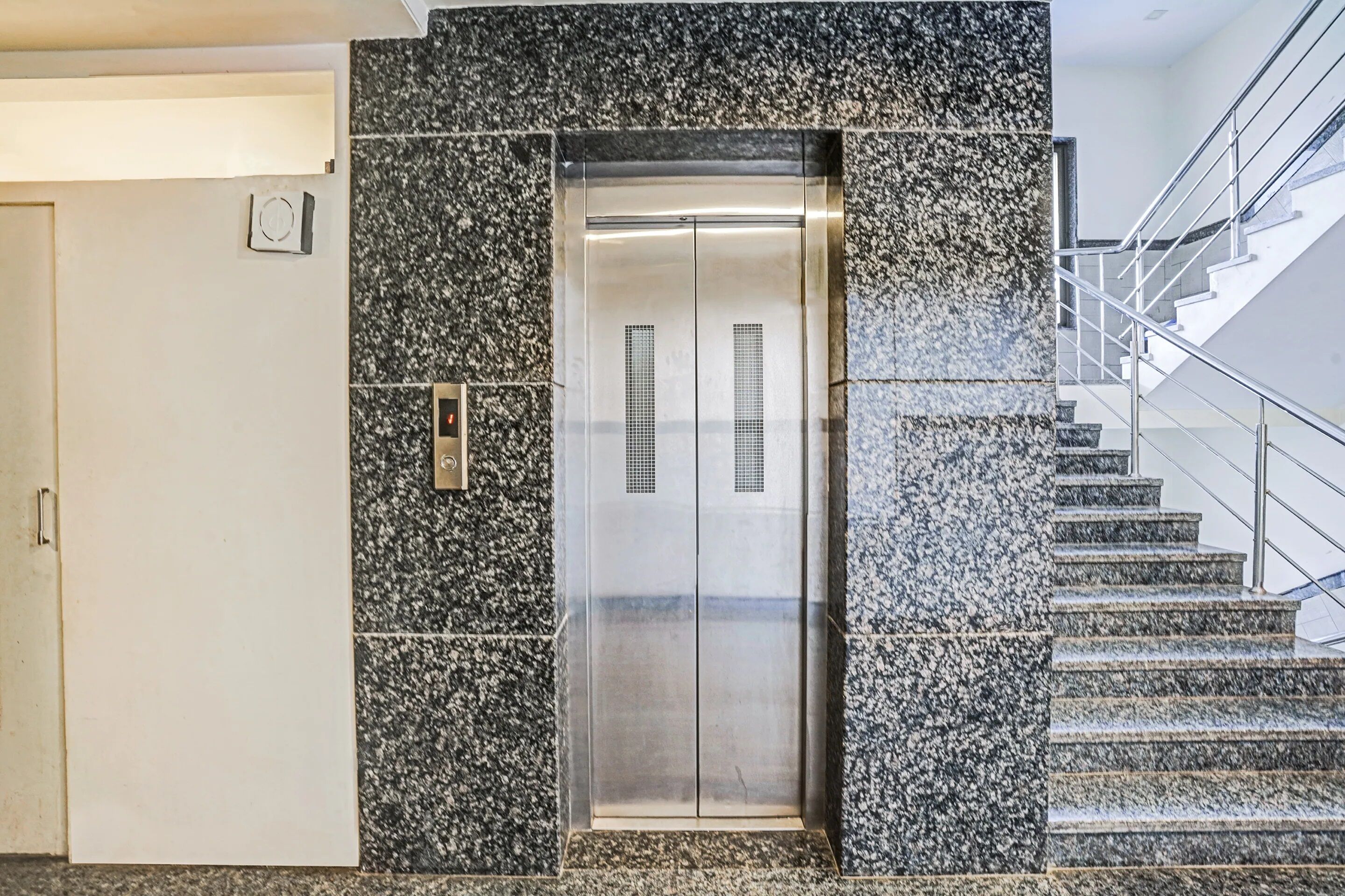 An elevator with a metallic finish, flanked by granite walls and a staircase with metal railings.