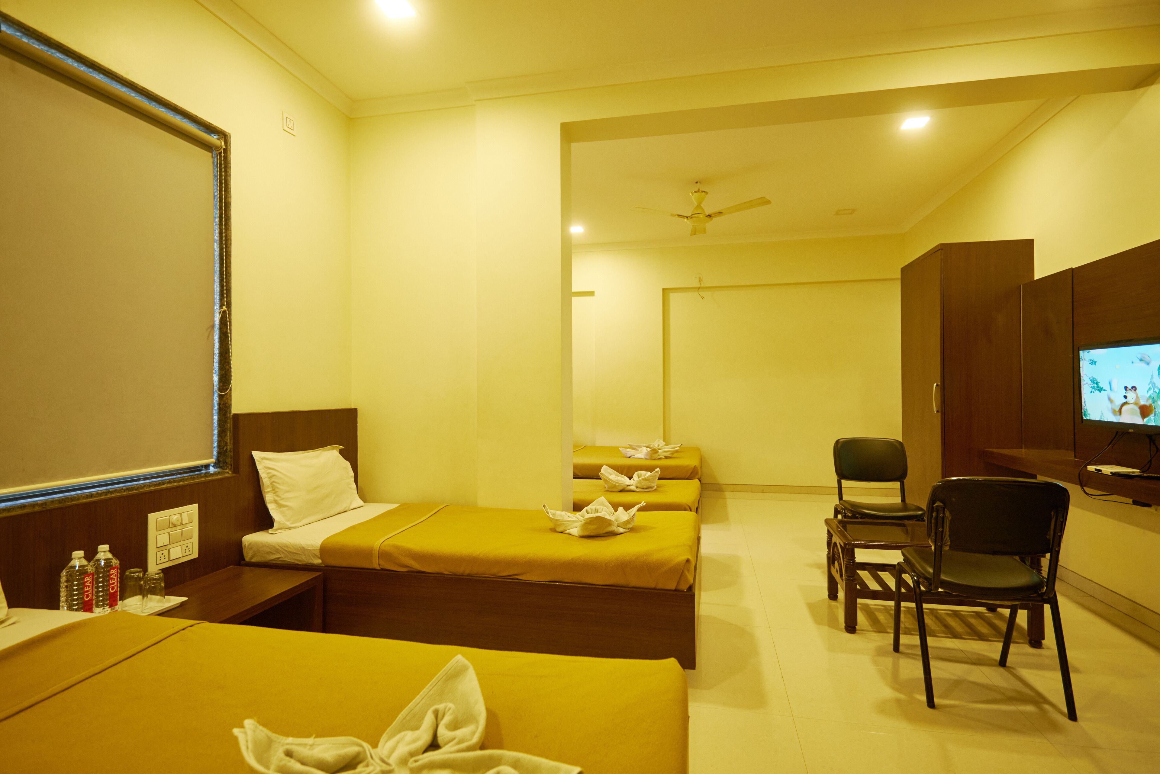 A hotel room with three single beds, a desk with a chair, a television, and a ceiling fan.