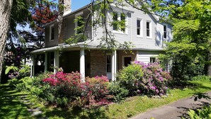 A two-story house with a stone chimney, a covered porch, and a garden with pink flowers. - Comfort, Charm and Tradition at once (Erdenheim)