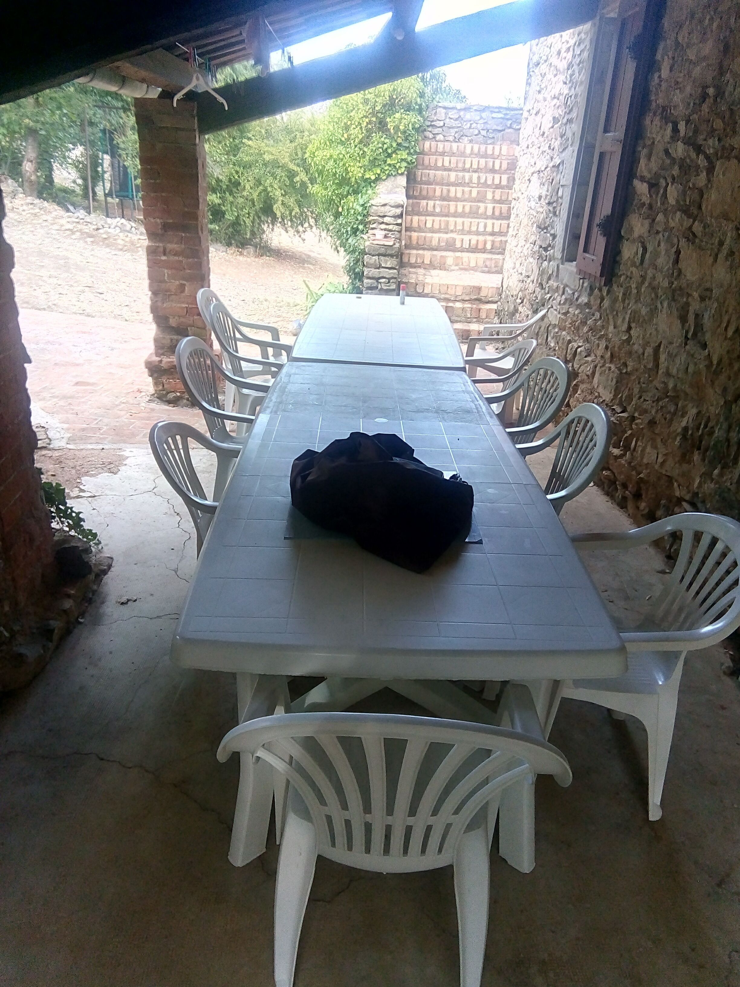 Un espace extérieur abrité, aménagé pour les repas, avec une longue table et des chaises en plastique blanches.