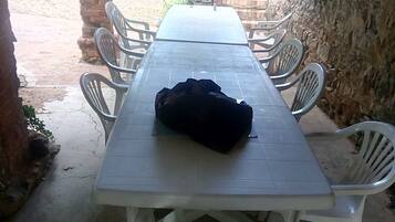 Un espace extérieur abrité, aménagé pour les repas, avec une longue table et des chaises en plastique blanches.
