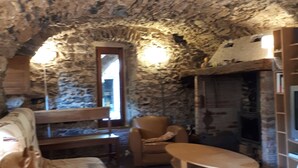 A rustic stone interior with a wooden sofa, a leather armchair, and a table. A fireplace is visible in the background.