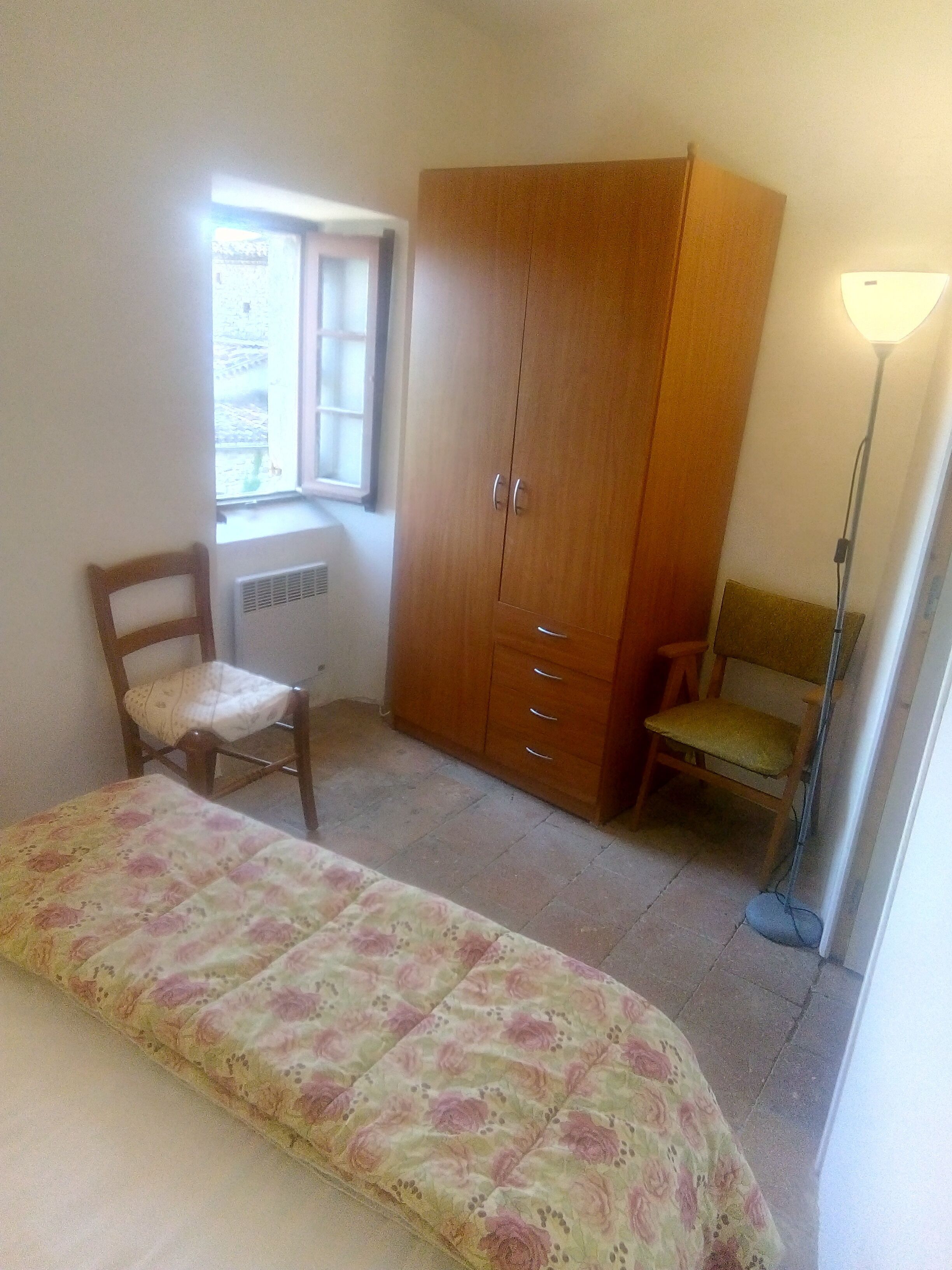 Un dormitorio con cama, armario de madera, silla, radiador y ventana.