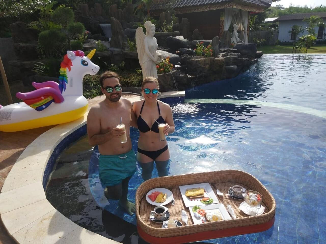A swimming pool with a small waterfall feature, a unicorn float, and a wicker table with food and drinks.