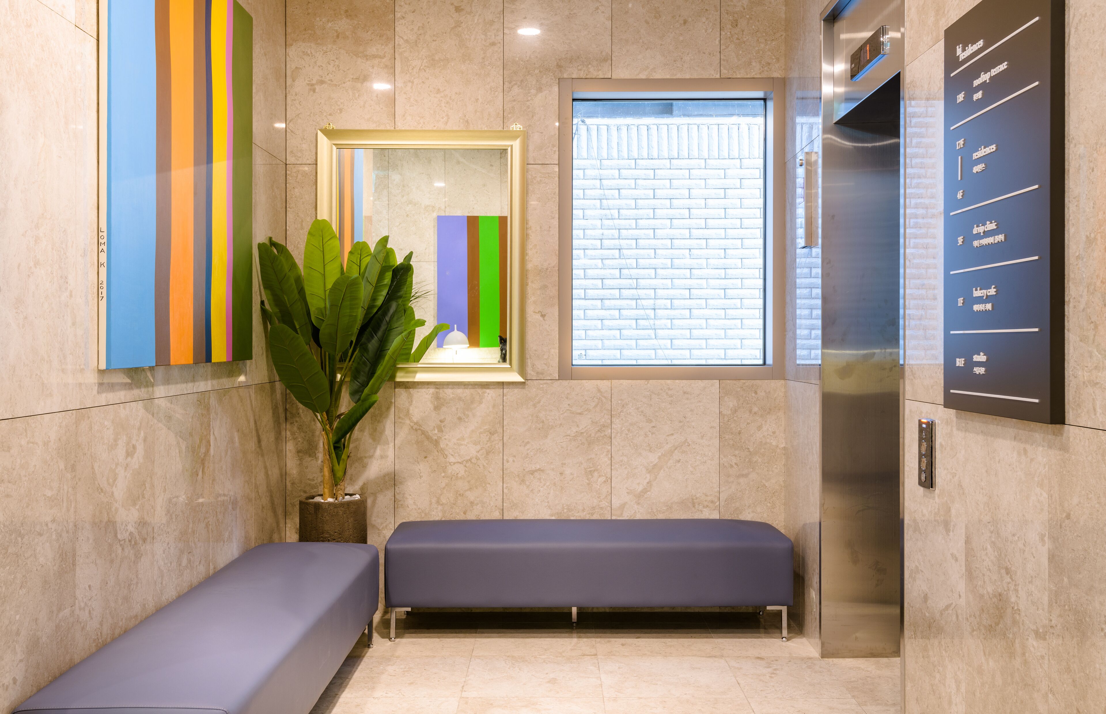 A waiting area with a bench, a potted plant, a colorful artwork, a window with a brick pattern, and an elevator with a digital display.