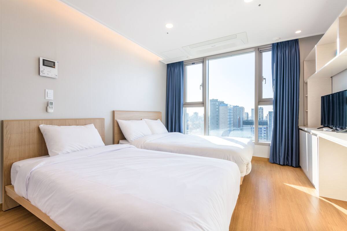 A hotel room with two beds, a desk, a TV, and a large window with curtains.