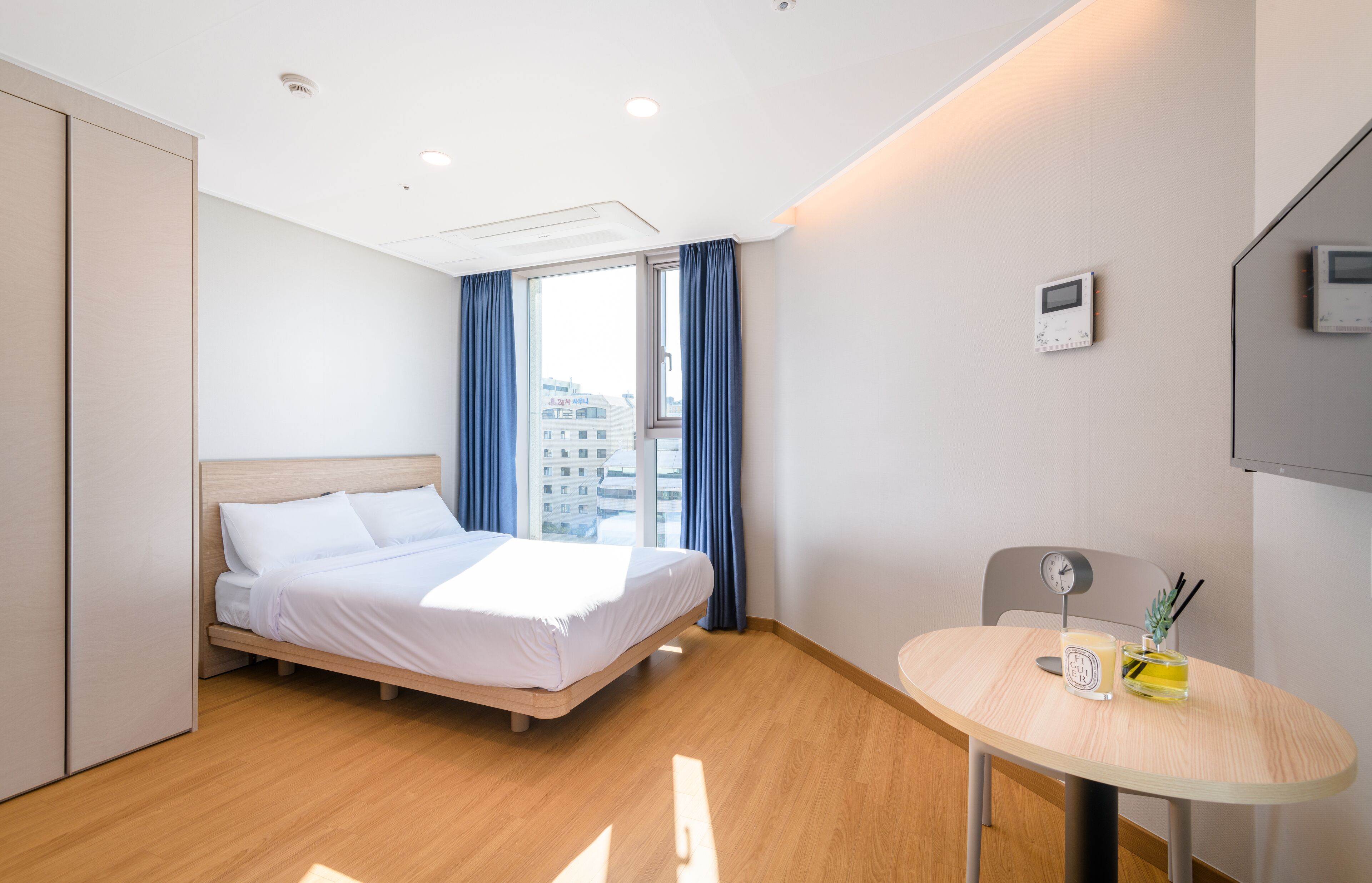 A modern hotel room with a bed, a small table with a clock and a bottle, and a window with curtains.
