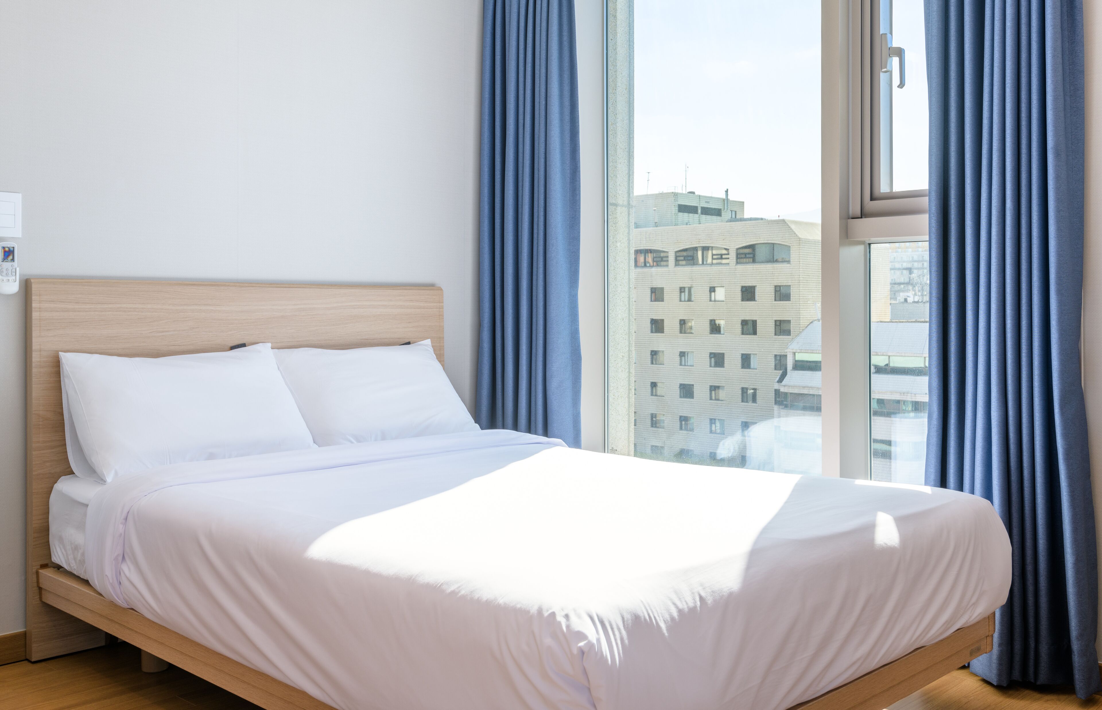 A neatly made bed with white linens in a hotel room with a large window and blue curtains.